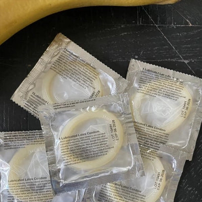 Several unopened lubricated latex condom packets arranged on a dark surface. The transparent wrappers show the circular condoms inside and feature printed text with product information and instructions.