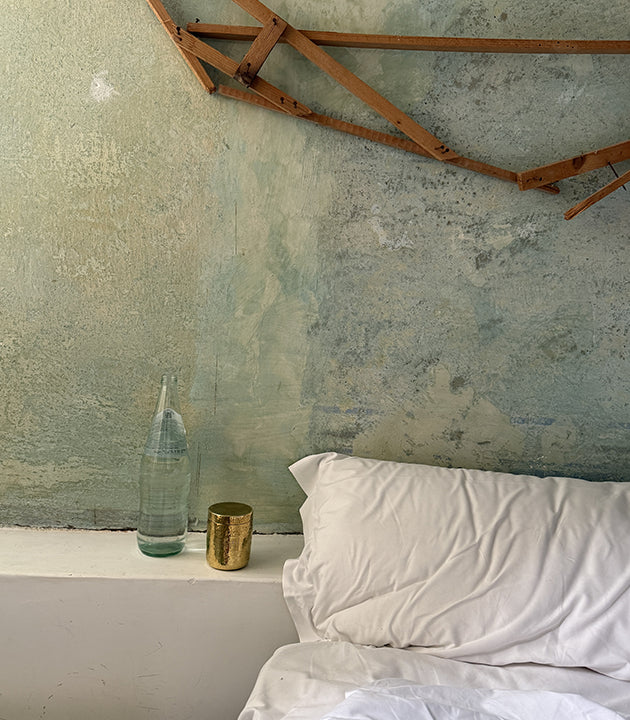 Meg Carozza's nightstand...Minimalist bedroom with rustic features showing a white bed with crisp white linens against a textured concrete or plaster wall with mottled green-gray patina. Wooden roof beams are visible at the top of the image. A clear glass water bottle and small brass cup sit on what appears to be a white bedside shelf. The space has a serene, wabi-sabi aesthetic with natural materials and subdued colors.