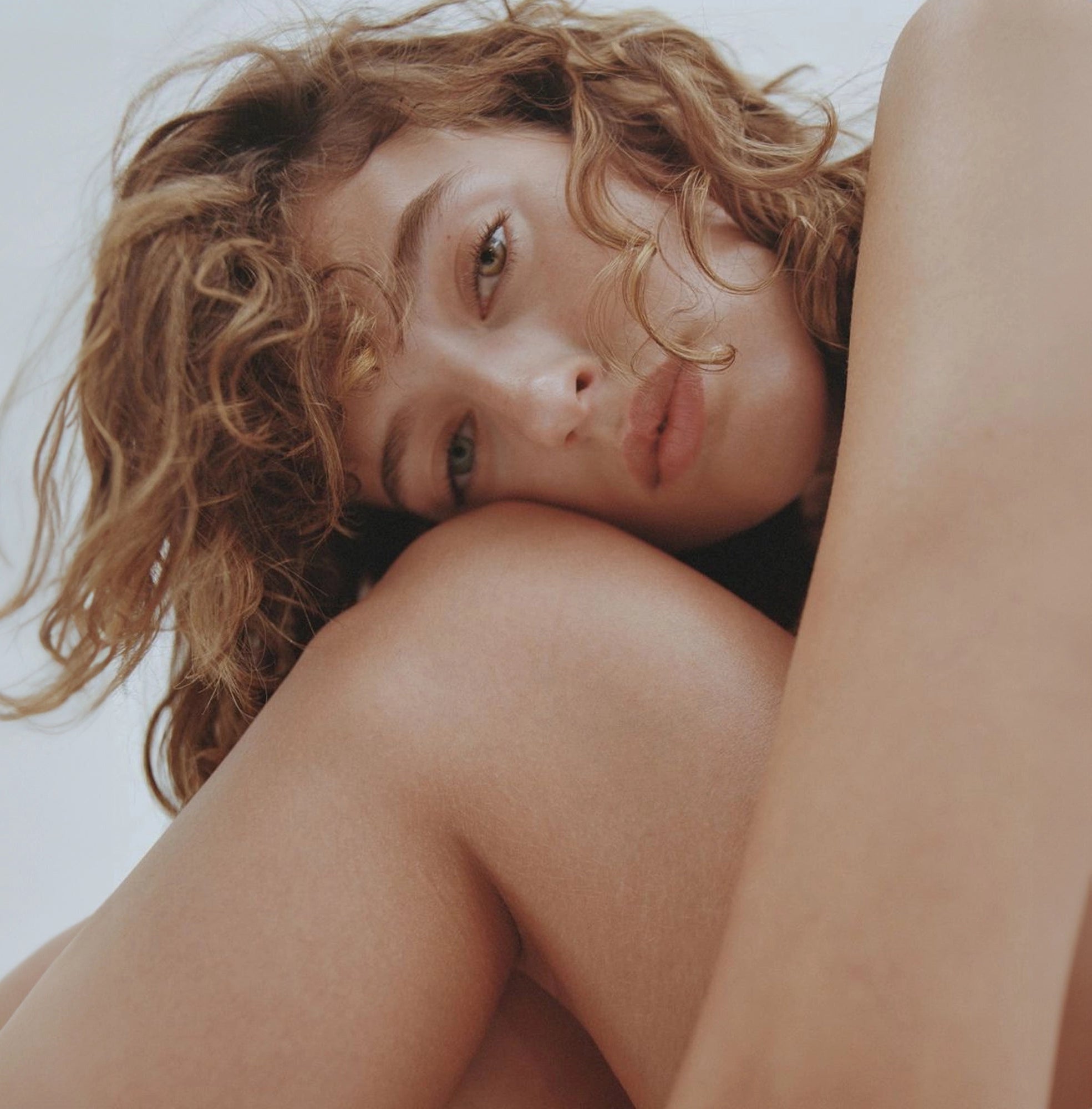 Close-up portrait of a woman with curly blonde hair and natural makeup resting her head on her arm, conveying a sense of comfort and intimacy in a minimalist setting.