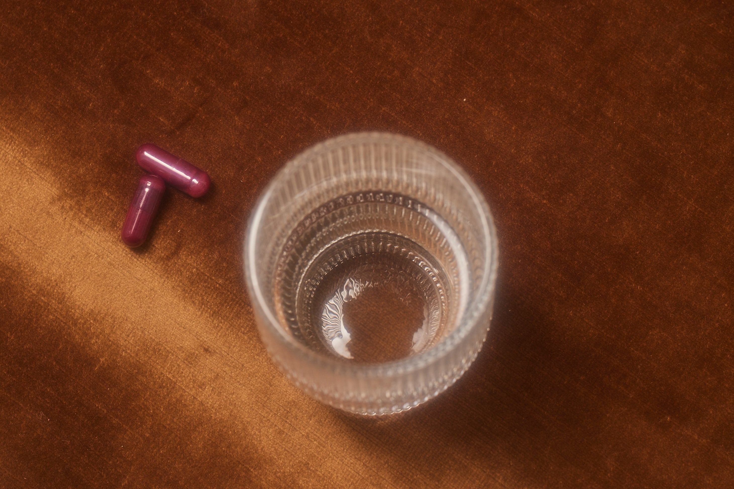 Overhead view of two dark purple capsule pills next to an empty glass drinking cup on a wooden surface. One pill has a small blue dot marking. The textured glass has ribbed sides and a small amount of liquid remaining at the bottom. The image shows what appears to be a supplement or medication ready to be taken with water.