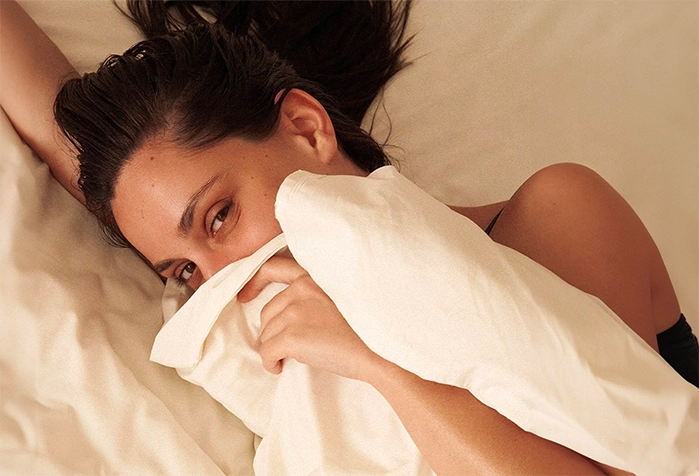 Woman with dark hair lying in bed, partially covering her face with a white sheet while gazing at the camera. The intimate, natural portrait conveys comfort and a relaxed morning aesthetic against cream-colored bedding.