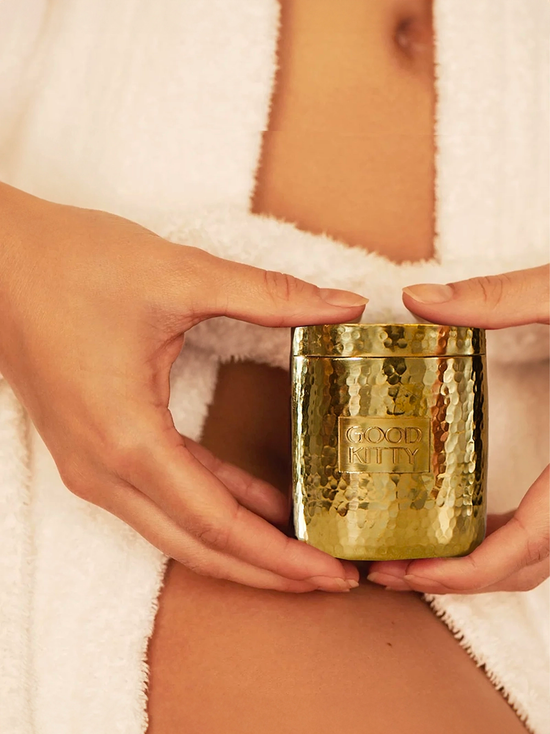 Hands cradling a hammered gold Good Kitty container against a white towel background, suggesting intimate wellness care. The word 'sustainable' appears vertically with a 'learn more' button below.