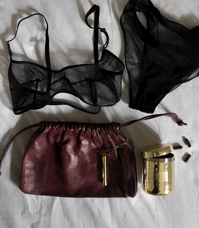 Styled flat lay featuring black sheer lingerie, a burgundy leather drawstring pouch, a hammered gold Good Kitty container, and several burgundy UTI biome Shield capsules arranged on white bedding. The composition suggests a premium, intimate wellness routine.