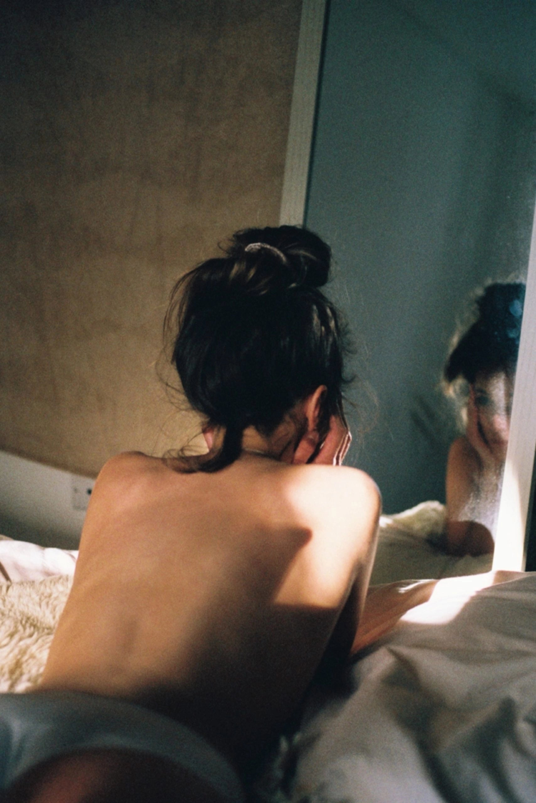 Woman with hair in a messy bun viewed from behind, sitting on a bed with soft white bedding and looking toward a mirror or window. The image captures natural light creating shadows across her bare back, conveying an intimate, contemplative morning moment in a bedroom with neutral-toned walls.