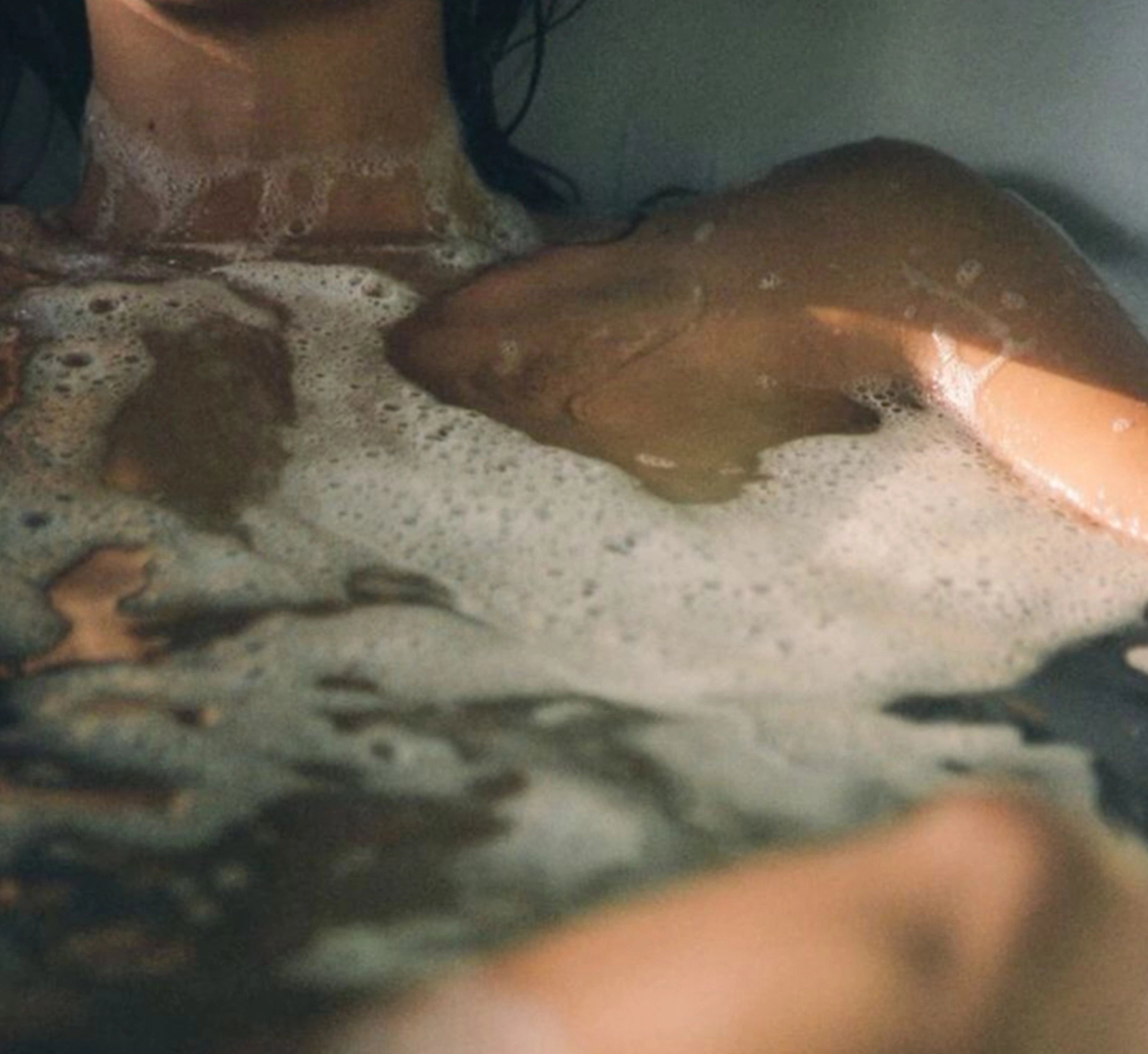Close-up artistic photograph of a person partially submerged in a bathtub with soapy bubbles and foam. The image shows the upper chest, neck, and shoulder area with skin visible through cloudy bathwater. The photograph has a soft, intimate aesthetic with muted colors and a dreamlike quality typical of film photography.