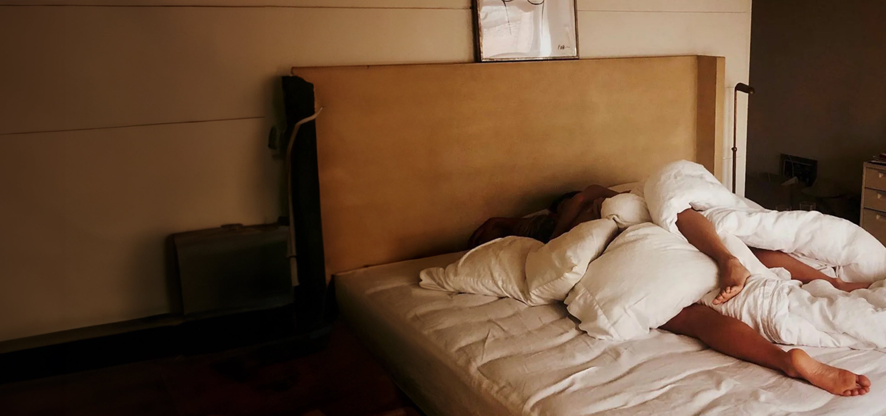 A dimly lit bedroom with warm amber tones showing a bed with white linens and a tall beige headboard against a light-colored wall. A person appears to be resting in the bed, partially covered by white bedding with one leg and arm visible but their face obscured by pillows or blankets. The room has a minimalist aesthetic with soft morning or evening light creating a serene, intimate atmosphere.