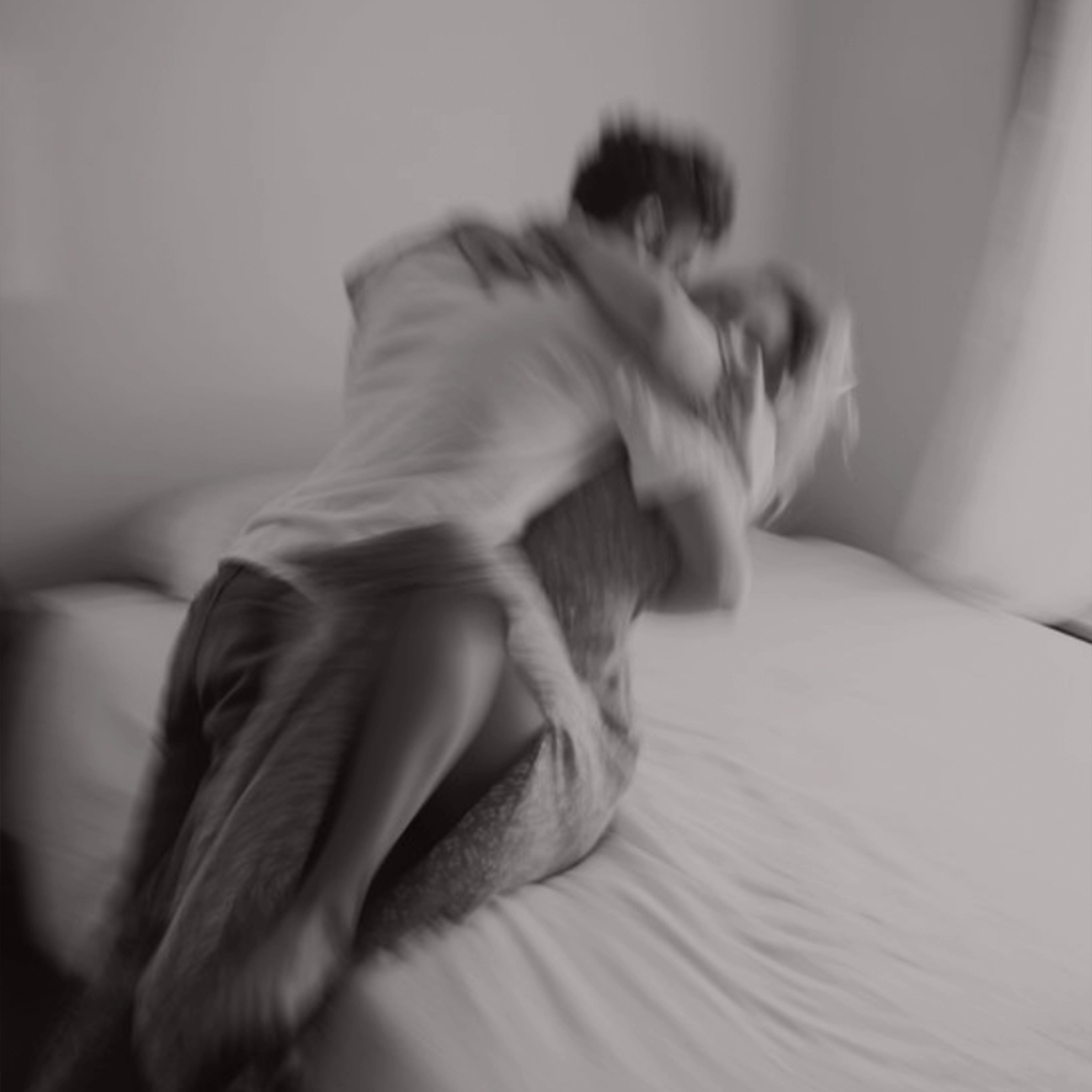 Black and white artistic photograph showing an intimate moment between a couple on a bed, captured with intentional motion blur. The image conveys intimacy and connection in a tasteful, non-explicit manner.