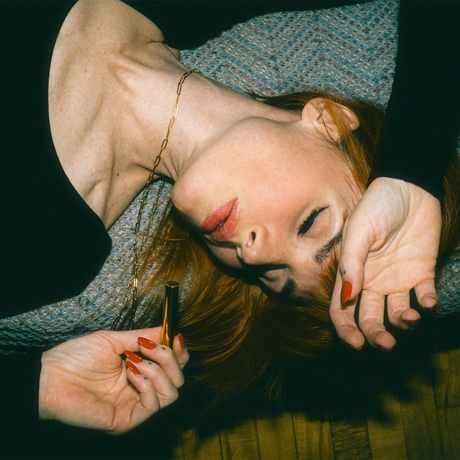 Artistic portrait of a person with reddish-brown hair lying on what appears to be a wooden floor. They're wearing a textured gray sweater and a delicate gold chain necklace. Their eyes are partially closed in a dreamy expression, and they have one hand near their face. The image has a moody, intimate quality with dramatic lighting against a dark background.