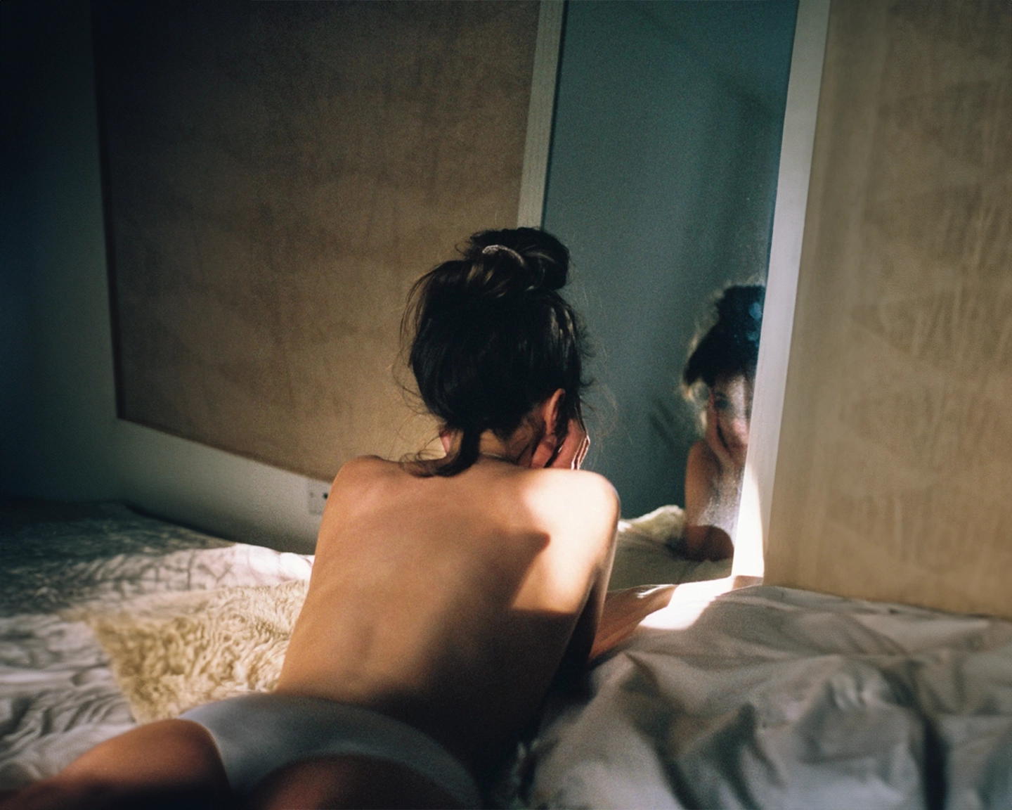 Back view of a person with dark hair in a messy bun sitting on a bed with rumpled white linens, looking toward a mirror or window. The person appears to be unclothed from the waist up, with their back to the camera. Soft, natural light filters into the room, creating an intimate, contemplative mood. The surrounding walls have a neutral, textured finish.
