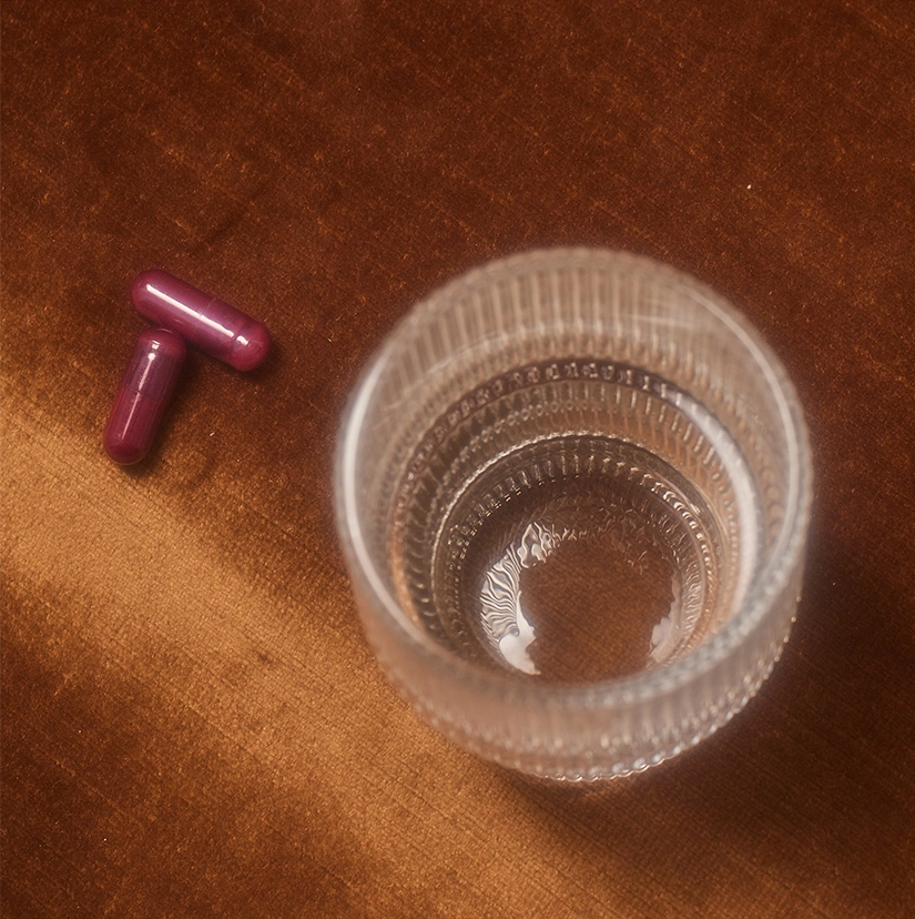 Overhead view of two burgundy UTI prevention capsules next to a textured glass of water on a wooden surface, displaying how the supplement is taken for intimate wellness.