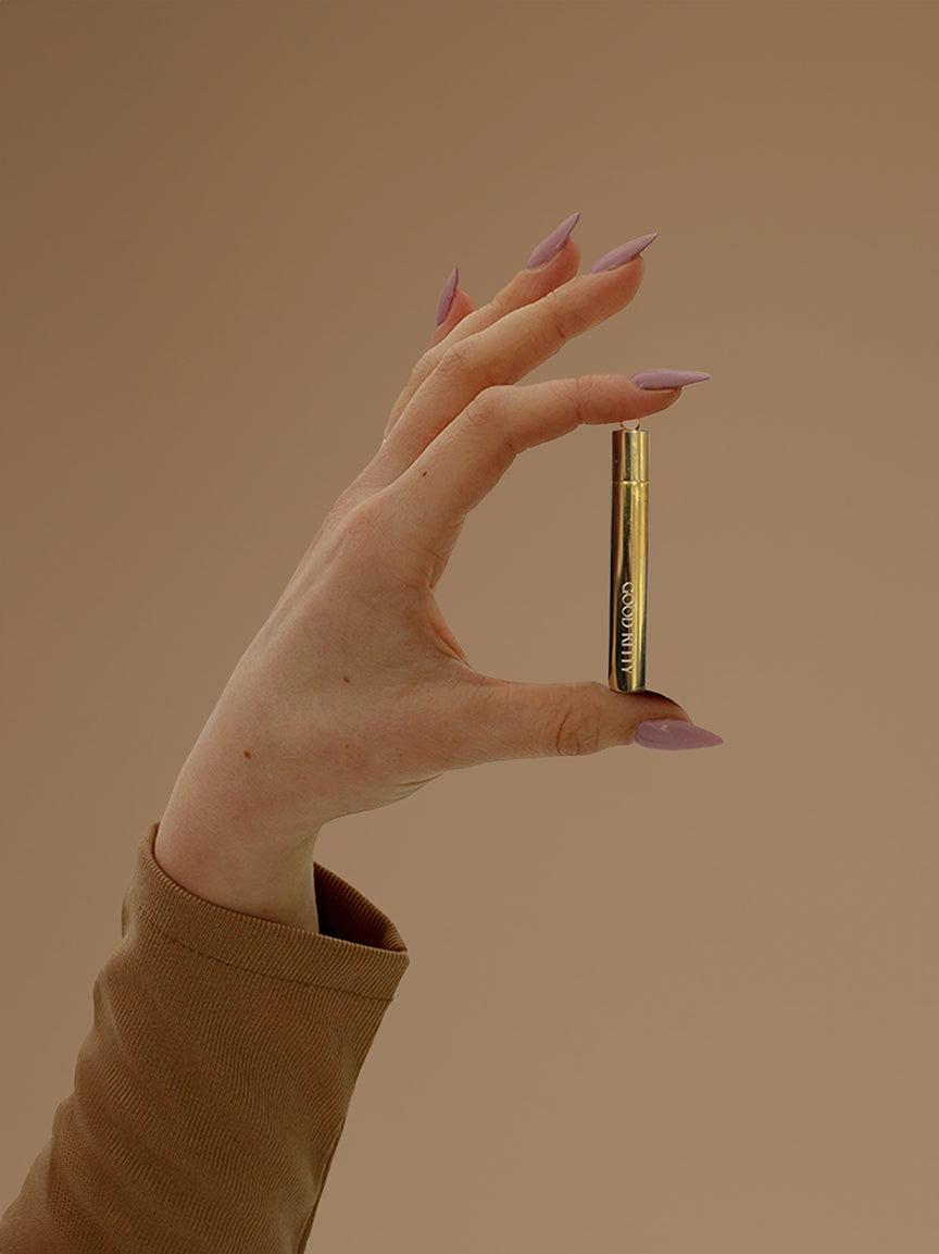 Close-up of a hand with long lavender-colored stiletto nails holding a gold cylindrical pendant or capsule against a neutral beige background. The hand emerges from a camel or tan-colored sleeve, and the gold tube appears to have some engraving or text on it. The minimal styling and soft tones create an elegant product showcase highlighting the jewelry piece.