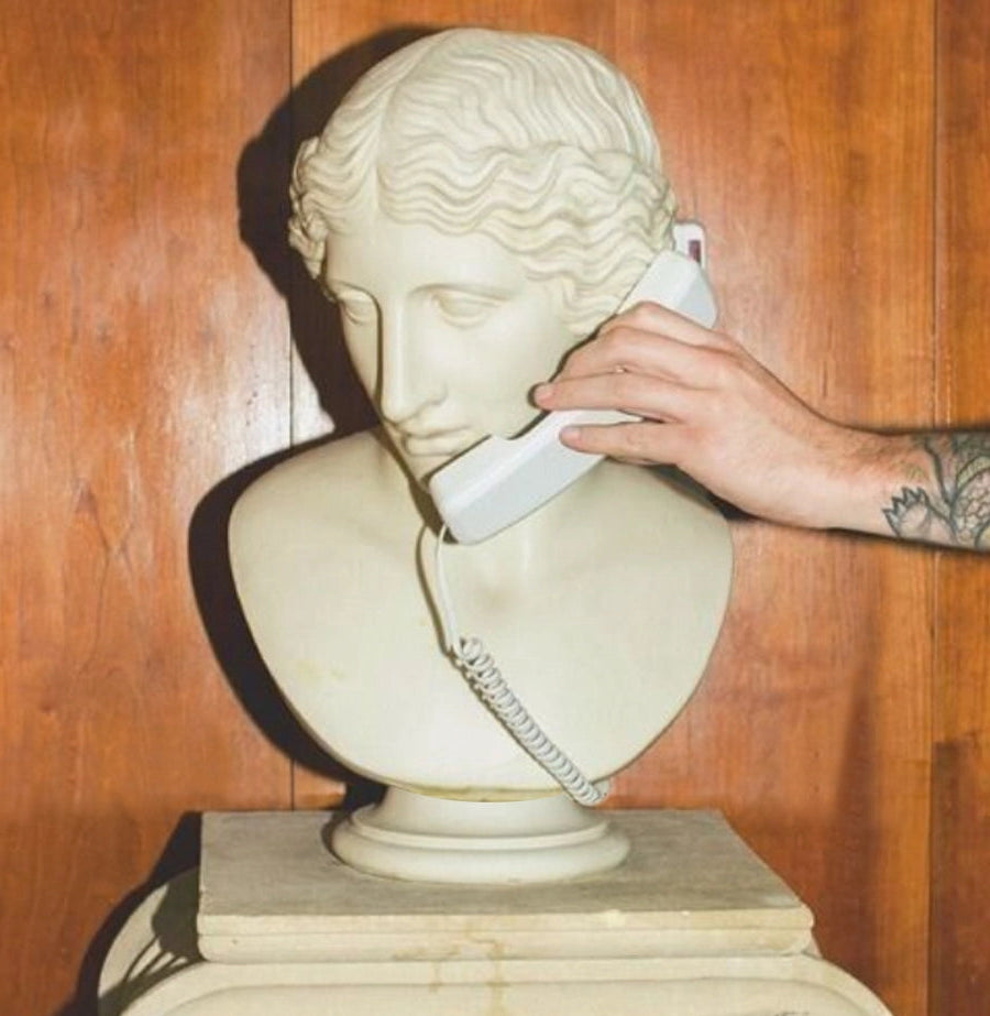 A white classical marble bust of a woman with wavy hair in Greco-Roman style posed with a corded telephone receiver held to its ear. A tattooed hand is placing the receiver against the side of the bust's head, creating a surreal, humorous juxtaposition between ancient art and modern technology. The sculpture is displayed on a pedestal against a wooden panel background.