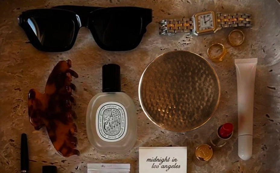 Flat lay of luxury personal accessories on a marble surface including black sunglasses, tortoiseshell hair clip, Diptyque perfume, gold hammered Good Kitty container, gold jewelry, watch, and cosmetics with 'midnight in los angeles' text card with 'uti biome shield $78" and a thumbnail image in the upper left corner.