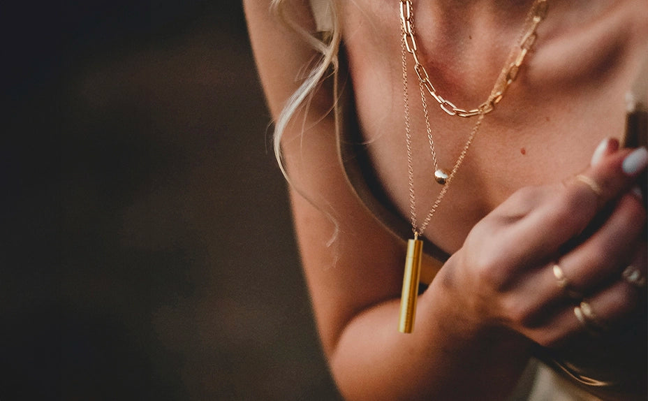 Close-up of a person wearing layered gold necklaces including a cylindrical pendant with product card showing 'the overnighter $49.00' and a hand holding the pendant in the foreground.