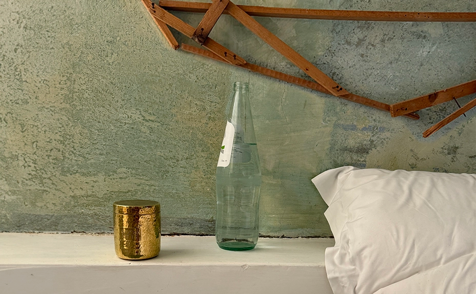 Bedroom still life featuring a hammered gold Good Kitty container, glass water bottle, and white pillow against a textured green wall with exposed wooden ceiling beams with product card showing 'uti biome shield $78" and a thumbnail image in the upper left corner.
