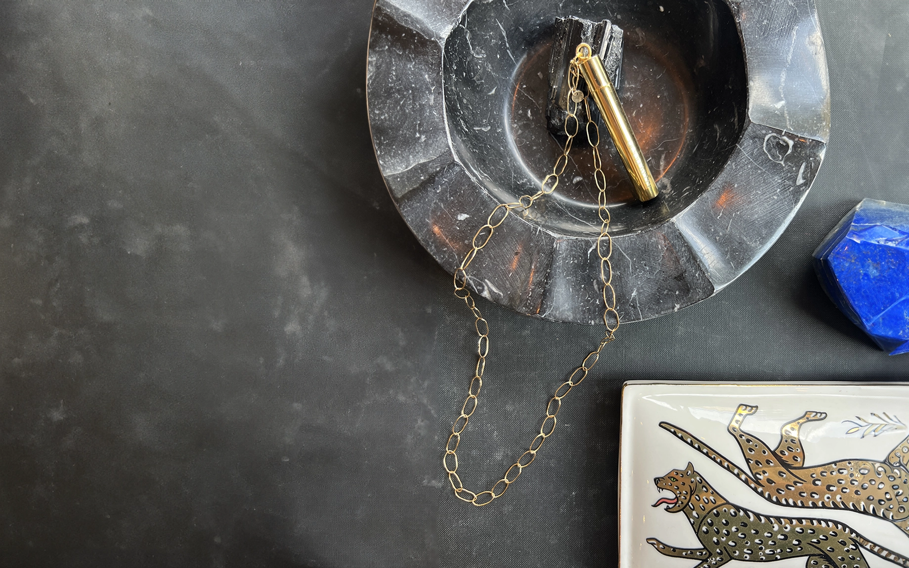 Elegantly styled photoshoot featuring luxurious 24k pendant designed by Meghan Carozza and chain by Mabel Chong for elevated Good Kitty UTI prevention and sexual wellness. Styled flat lay featuring marble, gold chain, and blue lapis against a dark background with text 'our ethos' and 'Female-led sustainability with purpose: packaging, collaborations, and initiatives that elevate women's lives.
