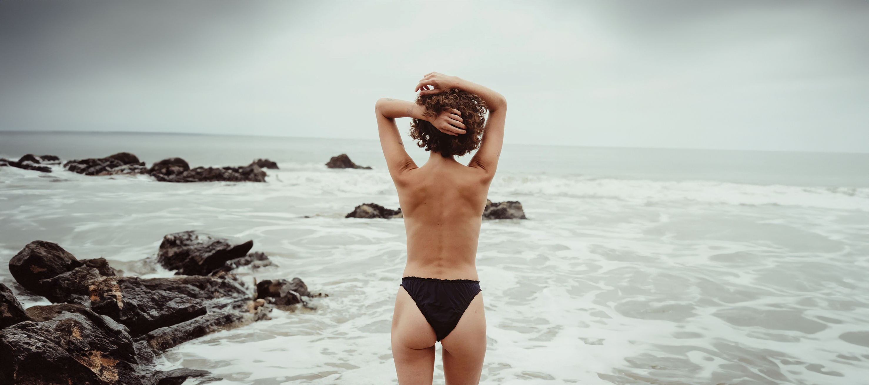 Woman standing in the ocean wearing black underwear with waves breaking around rocks.