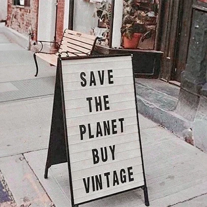 A-frame sidewalk sign outside a vintage store that reads 'SAVE THE PLANET BUY VINTAGE' in black text on a white letterboard. The sign is placed on a sidewalk in front of a storefront with plants visible in the window.