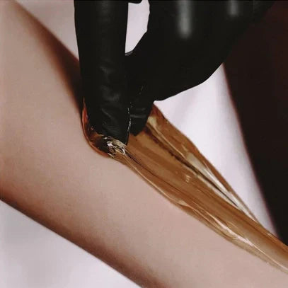 Close-up photograph of golden-brown liquid being poured from a black container, creating a smooth stream against a white background. The image captures the viscous texture and amber color of what appears to be honey, syrup, or a similar substance.