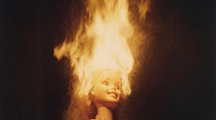 A doll or mannequin head with long blonde hair on a black background, engulfed in a large flame effect. No visible brand text or logos.