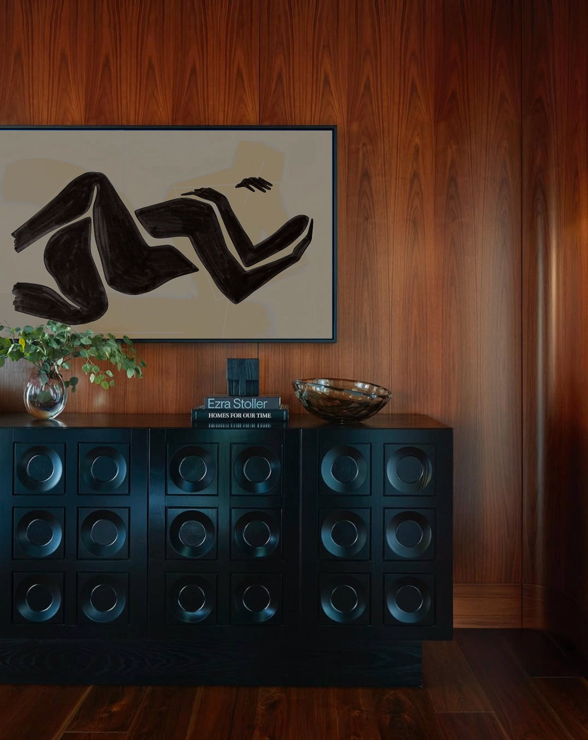 Stylish interior design featuring a dark textured cabinet with circular indented panels against rich wooden wall paneling. Above the cabinet hangs a minimalist black and white abstract art piece by Astrid Babayon that Meg Carozza blew up onto a canvas. Mid century cabinet, moody interior, wood clad walls.