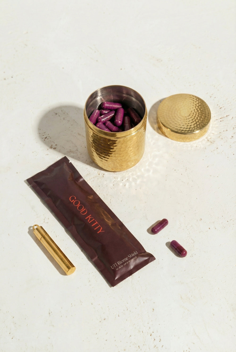 Gold container with purple capsules, brown packet labeled 'GOOD KITTY', and gold tube on a light surface.