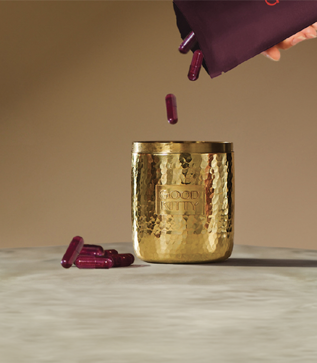 Burgundy UTI biome Shield capsules being poured from a purple pouch into a hammered gold vessel with the Good Kitty logo. Several capsules rest on a marble surface beside the gold container.