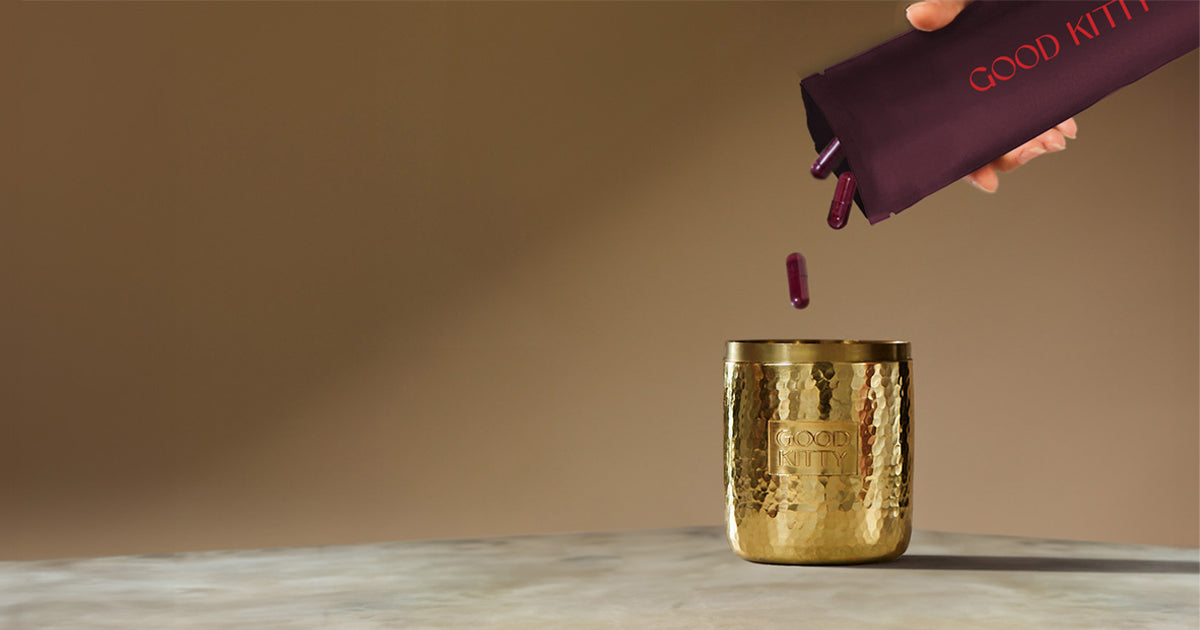 Burgundy UTI biome Shield capsules being poured from a purple pouch into a hammered gold vessel with the Good Kitty logo. Several capsules rest on a marble surface beside the gold container.