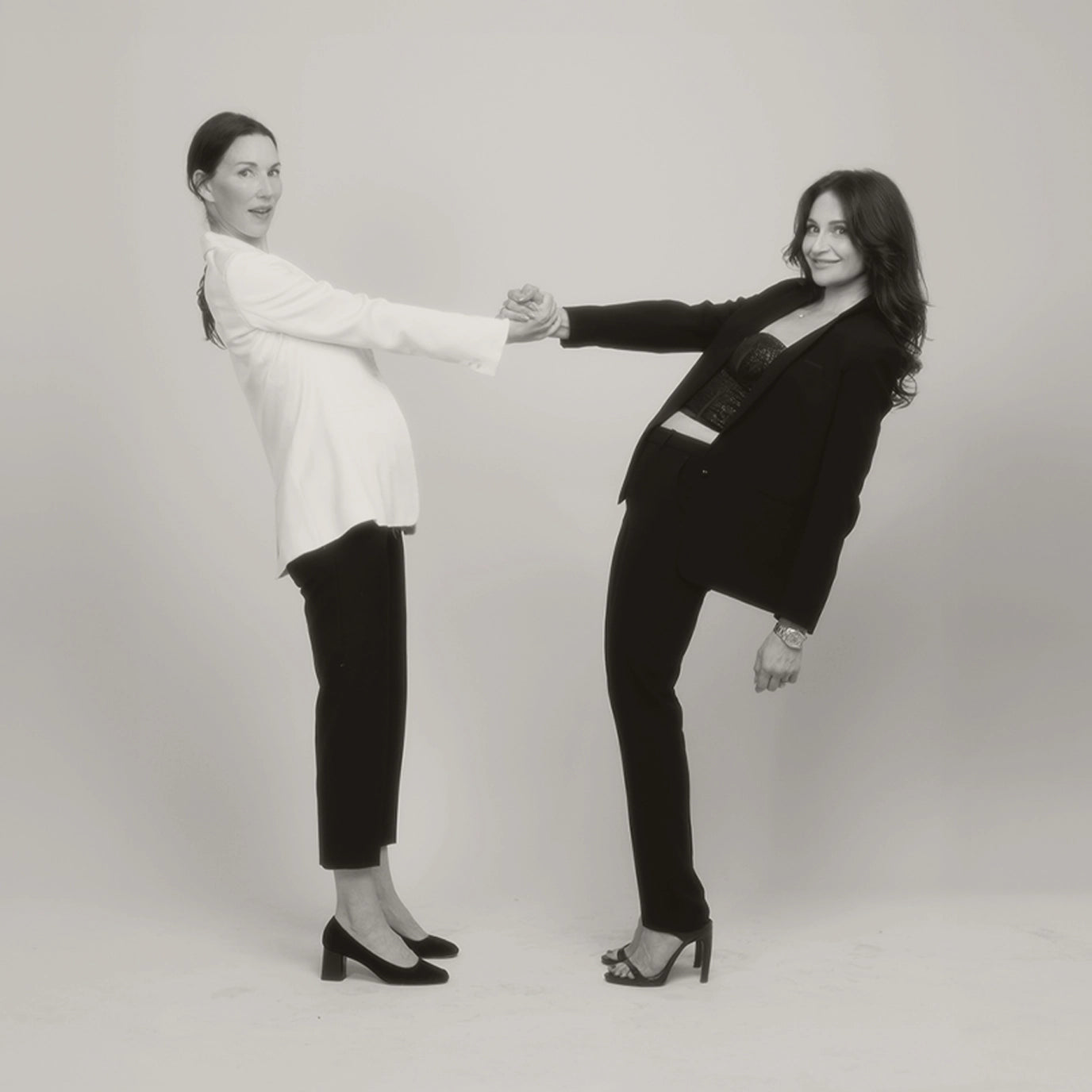 Two women holding hands and leaning back against a plain background.