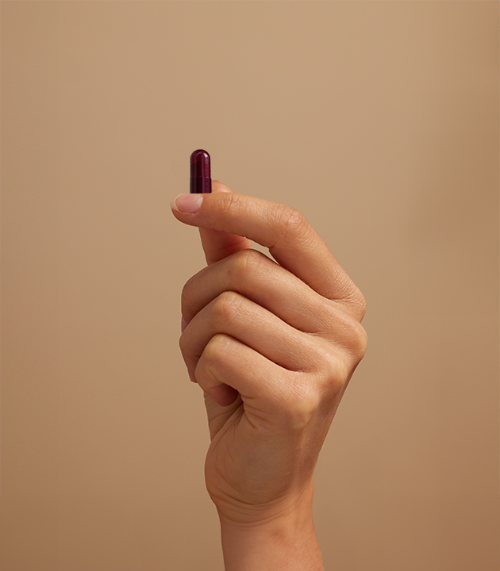 This image shows a hand holding UTI biome Shield capsule or pill between the thumb and index finger. The background is a neutral beige color, creating a minimalist, clean presentation.
